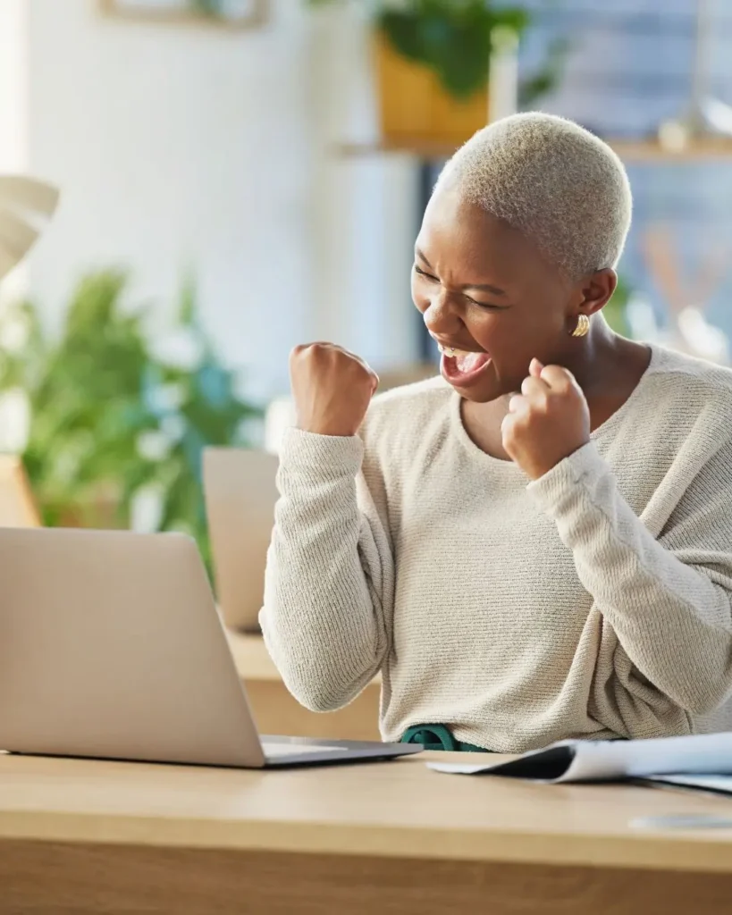 A woman sitting in front of her laptop with her arms raised in a pose of victory and joy, celebrating a breakthrough and growth.